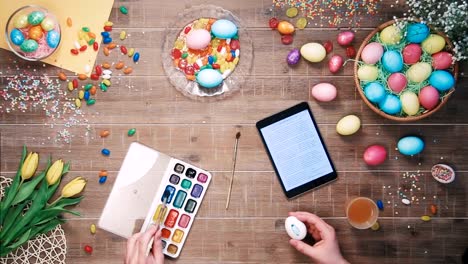 Man-painting-easter-egg-and-digital-tablet-lies-on-table-decorated-with-easter-eggs-Top-view