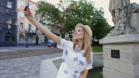 Niña-tomando-selfie-con-smartphone-sentado-en-la-fuente-en-la-ciudad.-Mujer-joven-feliz-sonriente-posando-para-el-retrato-del-uno-mismo-usando-la-cámara-del-teléfono-inteligente-iPhone.-Turismo,-instagram,-la-tecnología-moderna.