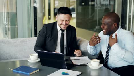 Two-businessmen-talking-ang-laughing-in-modern-cafe.-Business-colleagues-having-fun-and-joking-looking-at-laptop-computer-during-coffee-break