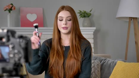 Young-red-haired-girl-blogger,-smiling,-talking-at-the-camera,-showing-a-new-purchase,-makeup-brush,-make-up-concept,-home-comfort-in-the-background.-60-fps