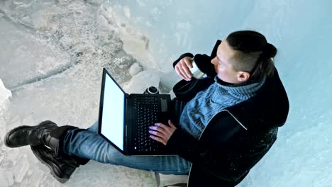 Man-is-sit-on-the-internet-in-laptop-in-an-ice-cave.-Around-the-mysterious-beautiful-ice-grotto.-User-communicates-in-social-networks.-He-crazy-strange-hipster-in-black-coat-and-fashionable-hairdo.