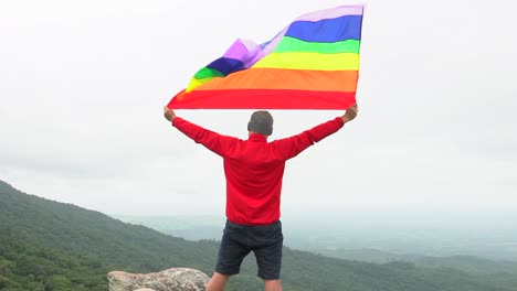 Mann-erhöhen-Regenbogen-Farbe-LGBTI-Fahnenschwingen-in-harten-Wind-auf-Mountain-Top-Sicht