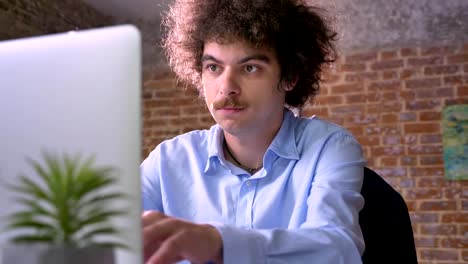 Serious-nerdy-businessman-with-curly-hair-typing-on-laptop-and-sitting-in-modern-office,-programmer-concept