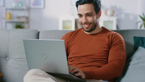 Alegre-joven-sentado-en-un-sofá-tiene-ordenador-portátil-en-su-regazo,-navega-a-través-de-Internet,-redes-sociales,-hace-compras.-Hombre-en-el-inicio-usando-Laptop-sentado-en-un-sofá.