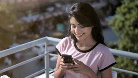 Eine-schöne-junge-Frau-lächelnd,-während-Sie-mit-dem-Handy-oder-Smartphone-sitzt-in-einem-Café-auf-der-Dachterrasse-vor-der-belebten-Straße.