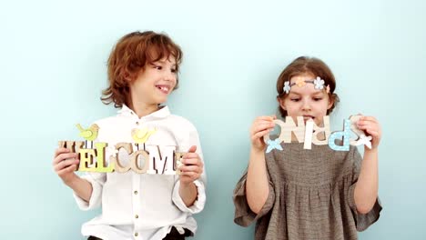 Studio-portrait-of-cute-school-children-with-wooden-signs.-Spring-holidays,-pastel-colors.-The-boy-is-wearing-a-white-shirt,-a-beige-dress-and-a-wreath-with-flowers