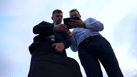 Two-young-businessmen-talking-and-using-tablet-pc-in-city.-Handsome-coworkers-discussing-and-working-on-digital-tablet-with-sky-at-background.-Colleagues-applying-mobile-technology.-Low-angle-view