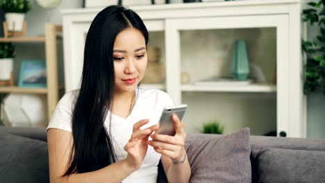 Cute-Asian-girl-is-using-smartphone-touching-screen-and-smiling-relaxing-on-sofa-at-home-alone-enjoying-modern-technology.-Youth-and-gadgets-concept.