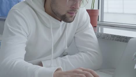 Portrait-of-a-young-stylish-handsome-bearded-guy-in-a-light-sweatshirt-and-typing-on-a-laptop