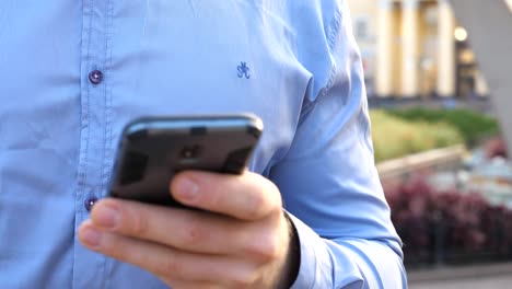 Nahe-Hand-von-jungen-Geschäftsmann-halten-und-berühren-Smartphone.-Unerkennbarer-Mann-steht-auf-der-städtischen-Straße-und-stöbert-Nachrichten-am-Telefon.-Guy,-der-Gadget-für-die-Arbeit-verwendet.-Langweilig-Dolly-erschossen