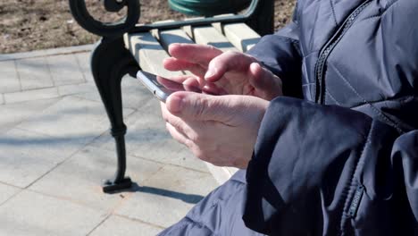 A-senior-elderly-woman-sitting-on-a-bench-in-park-outdoors-and-using-tapping-smartphone--she-is-useing-smartphone-to-connect-with-people-on-social,-texting-messages-ang-searching-internet