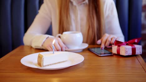 Close-up-of-waitress-serving-cake-with-burning-candle-for-woman-having-tea-and-browsing-smartphone-alone-in-cafe.-Birthday-gift-box-placed-on-table