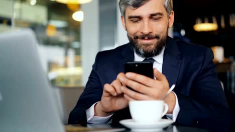 Bearded-office-worker-in-suit-using-smartphone-during-coffee-break-in-cafe