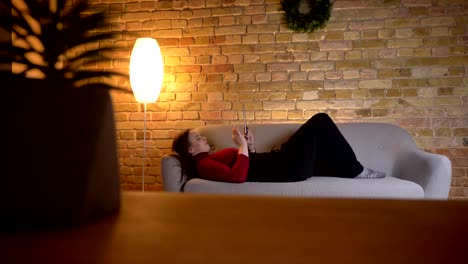 Closeup-shoot-of-young-pretty-caucasian-female-using-the-tablet-while-lying-on-the-sofa-in-a-cozy-apartment