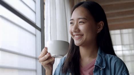 Close-up-attractive-portrait-smiling-young-asian-woman-holding-coffee-cup-standing-beside-window-at-home-office.