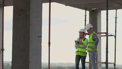 Trabajadorde-la-construcción-hombre-y-arquitecto-mujer-en-un-casco,-discutir-el-plan-de-construcción-de-la-casa,-hablarse-unos-a-otros-sobre-el-diseño,-sosteniendo-una-tableta,-mirar-los-dibujos,-fondo-de-los-rayos-del-sol