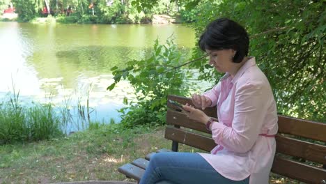 Eine-brünette-Frau-in-einem-Park-auf-einer-Bank-schaut-auf-ein-Foto-auf-ihrem-Smartphone