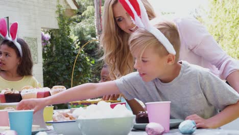 Group-of-children-wearing-bunny-ears-sitting-at-table-outdoors-enjoying-Easter-party-with-parents---shot-in-slow-motion