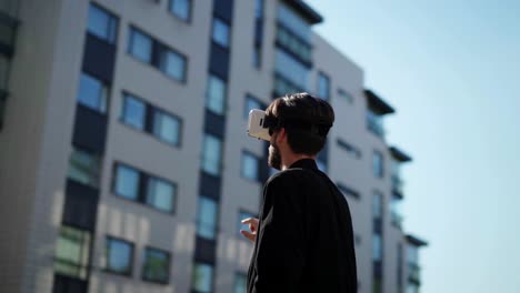 Panning-slow-motion-shot-of-young-man-wearing-vr-headset-standing-outdoors-in-city-street-and-exploring-data-in-sunlight