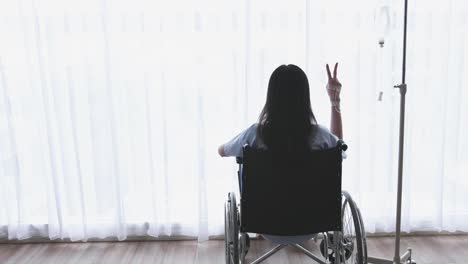 Asian-women-patient,-disabled-leg-sitting-in-a-wheelchair-and-showing--encouragement-symbol-with-two-fingers.