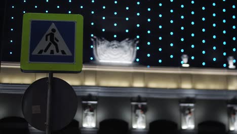 Close-up-view-of-pedestrian-crossing-sign-against-the-modern-shopping-center-with-the-blinking-night-illumination-and-plaster-statues-along-the-wall-at-night.-Stock-footage.-Beautiful-urban-buildings