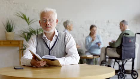 Rack-focus-of-three-senior-people,-two-elderly-women-and-disabled-old-man-in-wheelchair,-playing-cards-in-nursing-home.-Intelligent-old-man-reading-book-and-looking-at-camera-on-foreground