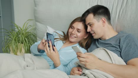 Atractivo-hombre-y-mujer-usando-teléfonos-inteligentes-en-la-cama-hablando-sonriendo-en-casa