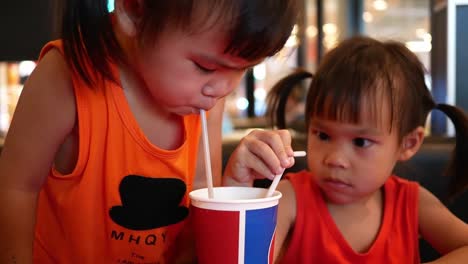 Pequeña-niña-asiática-con-su-hermana-bebiendo-su-bebida-juntos-mientras-se-sienta-a-esperar-comida-en-el-restaurante.-Enfoque-selectivo.