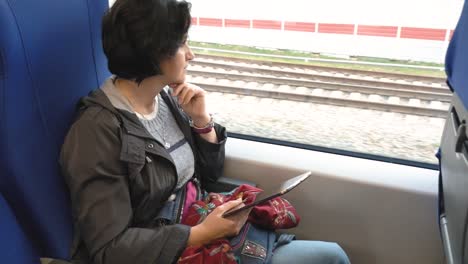 caucasian-woman-sits-train-window-holds-tablet-looks-out-window-props-her-chin