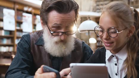 Intelligenter-Senior-Mit-zerknitterter-Gesicht-und-grauem-Bart-zeigt-etwas-auf-seinem-Handy-zu-seiner-lächelnden-Teenager-Enkelin,-während-sie-in-der-Bibliothek-sitzen