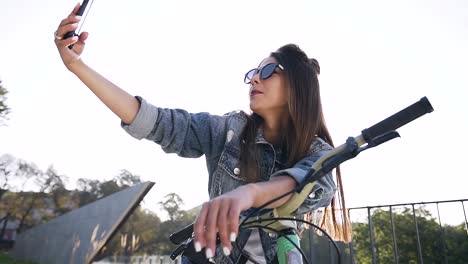 Wunderschöne-fröhliche-Mädchen-in-modischen-Sonnenbrille-und-stilvolle-Kleidung-sitzen-auf-dem-Fahrrad-und-machen-lustige-Selfie-unter-Sonnenlicht