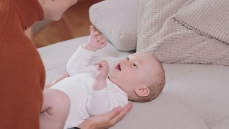 Nahaufnahme-des-Babys-auf-dem-Rücken-auf-der-Couch-liegend,-lächelnd-und-glücklich,-Hände-schüttelnd,-blickend-auf-seine-Mutter,-die-beim-Baby-kniet,-spricht-mit-ihm-und-lächelt