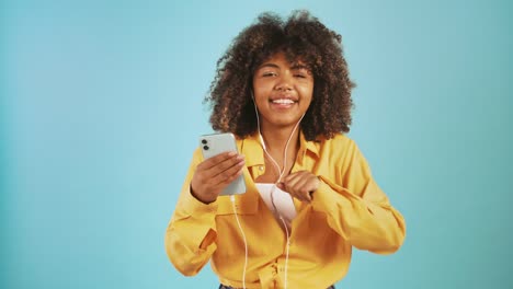 Dark-skinned-woman-listening-to-music-through-earphones-connected-to-smartphone,-dancing-and-smiling-posing-on-blue-background