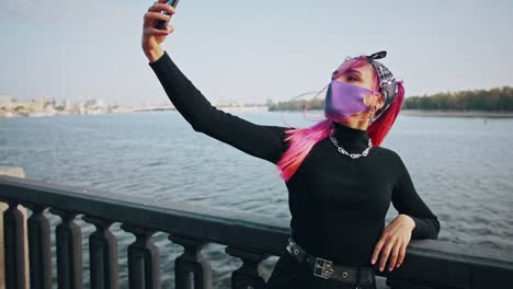 Hipster-female-in-informal-clothes-and-protective-mask-is-taking-selfie-on-smartphone,-leaning-on-fence-of-embankment