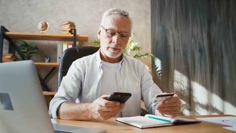 Senior-Mann-hält-Kreditkarte-und-geben-ihre-Nummer-in-Handy,-Einkaufen-online-arbeiten-im-Home-Office