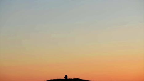 Man-hugs-woman-standing-on-top-of-hill-with-a-lot-of-sky-seen-far-away-at-sunset-sky-background-wide-shot.-Man-taking-picture-with-smartphone-of-view-from-mountain-at-honeymoon-holiday