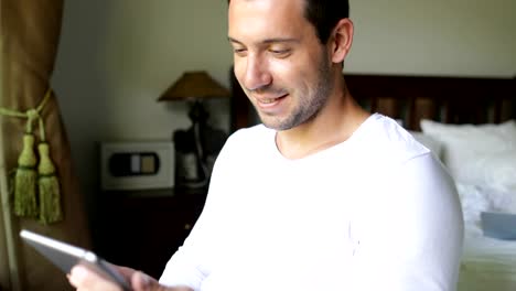 Young-Man-Using-Tablet-Computer-Drink-Coffee-Smiling-Guy-Chatting-Online-In-Bedroom