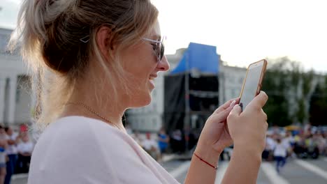 smiling-female-in-Sunglasses-writes-message-on-mobile-phone-standing-at-downtown