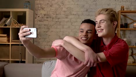 Two-international-gay-friends,-sitting-on-the-couch,-hugging,-making-selfies,-laughing.-Homeliness,-romantic-evening,-background,-hugs,-happy-LGBT-family-concept.-60-fps