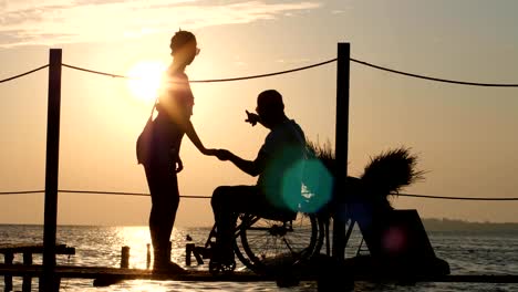 Silhouette-der-behinderte-Mensch-mit-Geliebte-Frau-auf-See-Pier-in-Abend-Abendrot-auf-Hintergrund-der-orangefarbenen-Himmel-und-Wasser