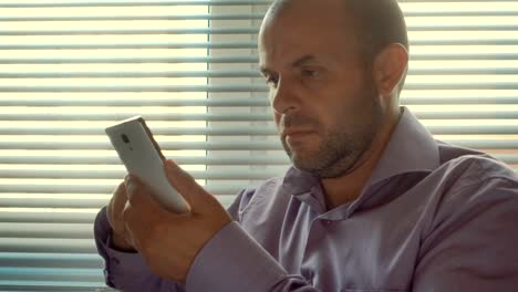 Young-businessman-standing-with-smartphone-by-window-in-the-office.-Close-up,-serious-man-sitting-by-out-the-window-and-uses-a-smartphone.-Thoughtful,angry,-problem,-social-network.