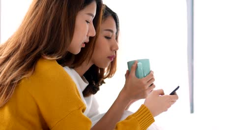 Beautiful-young-asian-lesbian-woman-couple-playing-mobile-phone-and-drinking-coffee-cup-on-bedroom-at-home.-Concept-lgbt.