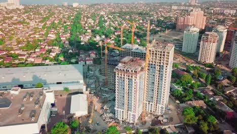 Aerial-drone-shot.-Construction-of-high-rise-buildings-in-the-developing-area-of-a-large-city.-Sunset-shot.-In-the-frame-moving-and-working-construction-cranes-and-many-houses-under-construction.-wide-shot