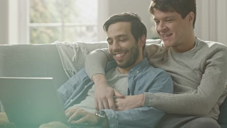 Dulce-pareja-gay-hombre-pasa-tiempo-en-casa.-Son-de-mentira-abajo-en-un-sofá-y-usar-la-Laptop.-Navegan-en-línea.-Mano-de-socio-es-alrededor-de-su-amante.-Sonreír-y-reír.-Habitación-tiene-un-Interior-moderno.