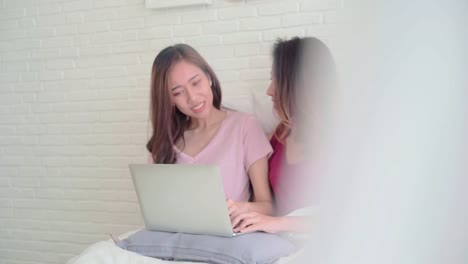 Young-Asian-women-lesbian-happy-couple-using-computer-laptop-checking-social-media-in-bedroom-at-home,-enjoy-love-moment-while-lying-on-bed-when-relaxed.-Lifestyle-LGBT-couple-together-indoors-concept