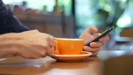 Primer-plano-de-la-mano-de-un-hombre-sosteniendo-y-usando-el-teléfono-inteligente-mientras-bebe-de-una-taza-de-color-naranja-de-humeante-café-en-una-cafetería-moderna-en-Medellín,-Colombia,-Latinoamérica.