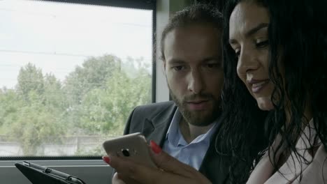 Closeup-of-young-business-partners-couple-sitting-in-a-moving-train-talking-and-analyzing-business-information-on-smartphone