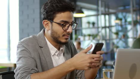 Hombre-árabe-guapo-usando-smartphone-y-sonriendo-a-la-cámara-en-el-café