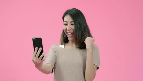 Junge-asiatische-Frau,-die-Smartphone-überprüft-Social-Media-Gefühl-glücklich-lächelnd-in-lässiger-Kleidung-über-rosa-Hintergrund-Studioaufnahme-erschossen.-Glücklich-lächelnde-entzückende-Frau-freut-sich-über-Erfolg.