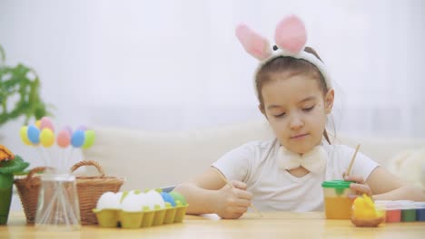 Little-cute-girl-is-having-fun-holding-a-paint-brush-in-her-left-hand.-Girl-with-a-beauty-spots-at-her-face-watching-at-nude-paint-brushes,-sitting-at-the-wooden-table-with-Easter-decorations.-Girl-is-amused-and-gives-up-holding-paint-brushes-and-hiding-h